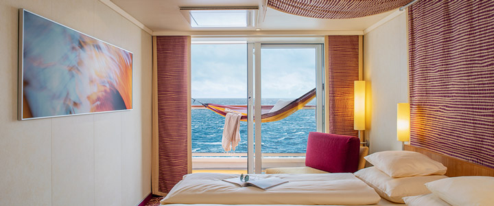 Blick durch eine elegante Außenkabine der AIDAsol auf den Balkon mit Hängematte und im HIntergrund das offene Meer.